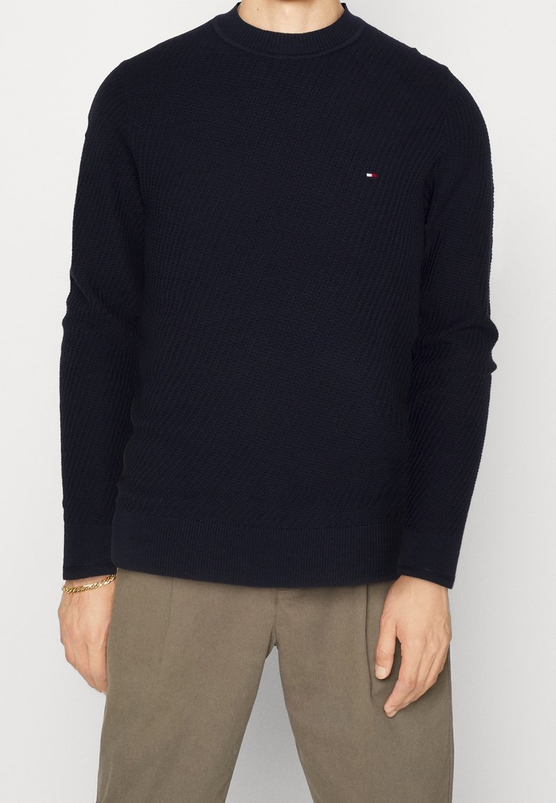 Homme portant un pull à manches longues noir texturé avec un petit logo sur la poitrine, accompagné d’un pantalon beige et d’un bracelet en or.