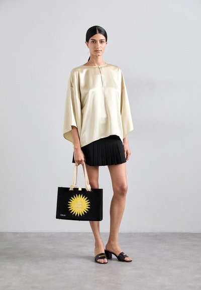 Cream satin top with a loose fit and a subtle neckline, paired with a pleated black skirt. The model carries a black tote with a yellow sun design.