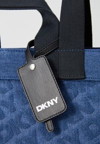 Mėlynas audinio pirkinių maišelis su tekstūrizuotu dizainu, juodomis rankenomis ir juodu odiniu žymeniu su baltu "DKNY" prekės ženklu.