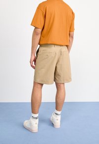 Mies, joka on pukeutunut beigeihin polvimittaisiin shortseihin, oranssiin t-paitaan, valkoisiin sukkiin mustilla raidoilla ja valkoisiin lenkkareihin, seisoo sinisellä lattialla.