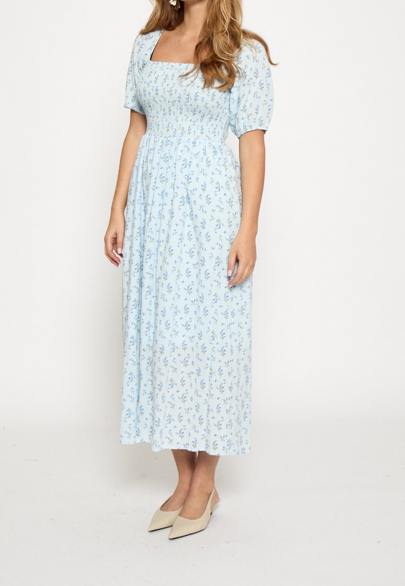 Femme portant une robe midi bleu clair avec de petits imprimés floraux, des manches ballons, un décolleté carré et des chaussures beiges à bout pointu.