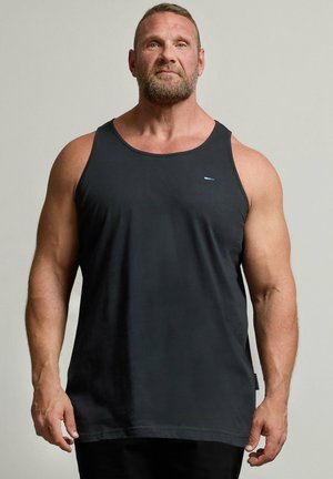 Spierbundige man met baard staat rechtop, draagt een mouwloos zwart hemd en zwarte broek, kijkt recht vooruit tegen een egale lichte achtergrond.