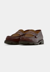 Mocassins de couro castanho com inserções de camurça e solas pretas robustas, exibidos lado a lado sobre um fundo branco liso.