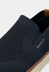Chaussure mule bleu marine avec texture en mesh sur le dessus, côtés élastiques et accents marron clair sur la semelle. Présente un logo de marque discret sur le côté.