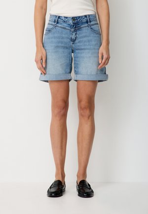 Woman wearing light blue rolled-up denim shorts, white top, and black loafers, standing against a plain white background.