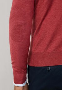 Zbliżenie ramienia osoby ubranego w czerwoną sweter z teksturowanym łokciem i niebieską koszulę z mankietami pod spodem.