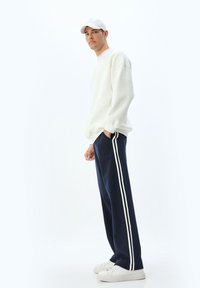 Sudadera blanca con hombros caídos, pantalones de chándal azul marino con franjas laterales blancas y zapatillas blancas. Diseño simple y moderno.
