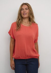 Haut corail ample avec des manches courtes, un col arrondi et un ourlet élastique froncé. Le tissu a une finition douce et texturée. Associé à un jean foncé.