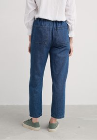 Person iført blå denimjeans med baglommer, hvid langærmet bluse og grønne sneakers, stående på en lys grå gulv.