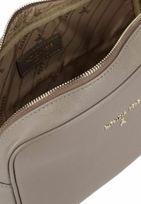 Bolso de cuero beige con superficie texturizada, cierre de cremallera y herrajes en tono dorado. El interior presenta un forro con estampado y etiqueta de la marca.