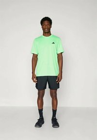 Camiseta atlética verde de manga corta con logo negro, combinada con pantalones cortos negros y zapatillas oscuras, sobre un fondo neutro.