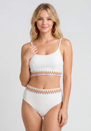Blondynka w białym, prążkowanym, dwuczęściowym stroju z kolorowym obszyciem w zygzaki, duże czerwono-złote efektowne kolczyki, uśmiecha się na szarym tle.