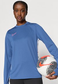 Μπλε αθλητική μπλούζα με μακρύ μανίκι και στενή σχεδίαση, με ροζ λογότυπο Nike στο στήθος. Κρατείτε μία ριγέ ποδοσφαιρική μπάλα σε άσπρο και μαύρο.