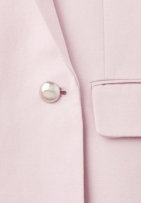 Primer plano de una chaqueta rosa claro que muestra un botón metálico brillante y una solapa de bolsillo parcialmente visible.
