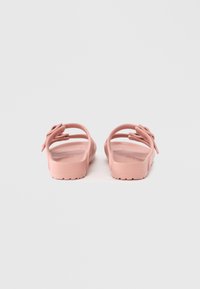 Rosa gummisandaler med flere tynne stropper og en strukturert såle. Bakfra vises en sløyfedetalj på siden. Lett design.