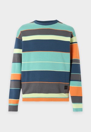 Jeune homme debout portant un pull rayé multicolore, un short en jean foncé jusqu'aux genoux, des chaussettes blanches et des baskets blanches, les mains dans les poches.