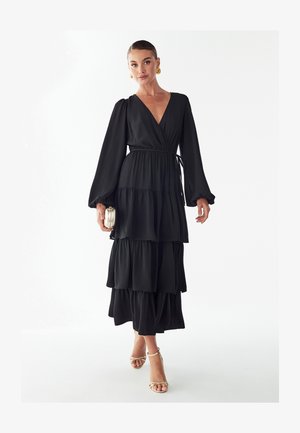 Sieviete, tērpusies garā melnā daudzpakāpju kleitā ar garām piedurknēm, zelta riņķa auskariem, zelta siksniņu papēžiem un turot rokās mazu zelta rokassomiņu.