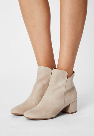 Beine in beigen Wildleder-Stiefeletten mit niedrigem Blockabsatz und leicht spitz zulaufender Spitze, stehend auf einer schlichten weißen Fläche.