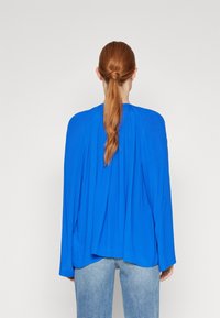 Blusa azzurra a maniche lunghe con pieghe, realizzata in tessuto leggero. Caratterizzata da una vestibilità comoda e scollatura rotonda, abbinata a jeans azzurri chiari.