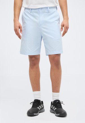 Άνδρας που φοράει ανοιχτό μπλε σορτς, άσπρες κάλτσες και μαύρα αθλητικά παπούτσια Adidas, στέκεται μπροστά σε ένα απλό λευκό φόντο.