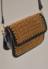 Rektangulær crossbody-taske med vævet front i strå og sider i sort læder. Har flaplukning og guldhardware-detaljer.
