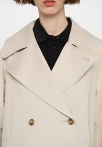 Beige double-breasted coat with large lapels, featuring two brown buttons and a subtle sheen. Black shirt with a ribbed texture underneath.
