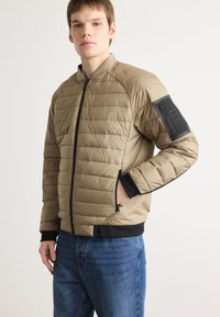 Casaco bomber acolchoado em caqui com design matelassé, punhos em malha pretos e bolsos laterais; possui um bolso com fecho no braço.