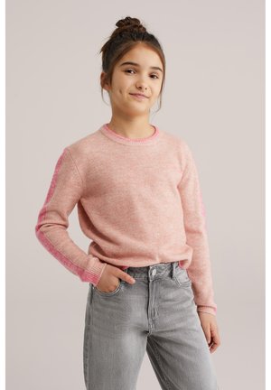 Noor tüdruk, kellel on kuklasse kogutud juuksed, seljas roosa kampsun ja hallid teksad, seisab ühe käega püksitaskus ühevärvilise tausta ees.