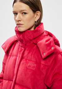 Sarkans, izmantojams pūšamais mētelis ar augsto apkakli un pogām ar spiedpogu. Mīksta, plīša tekstūra ar izšūtu logotipu uz krūtīm.