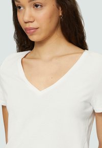 T-shirt en coton blanc avec décolleté en V, manches courtes et coupe ajustée. Texture lisse sans motifs ni graphismes visibles.