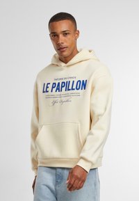 Crème hoodie met een voorzak, afgewerkt met blauwe tekst "LE PAPILLON" en "THÉORIE DU CHAOS." Zacht materiaal, standaard pasvorm en verstelbare capuchon.