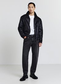 Uomo in piedi con le mani in tasca, indossando una giacca in shearling nera, una maglietta bianca, jeans scuri, una cintura nera e stivali eleganti neri su uno sfondo semplice.