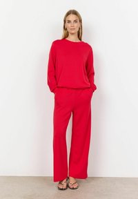 Piros, hosszú ujjú felső széles szárú nadrággal párosítva, mindkettő sima anyagból készült. Az outfit laza fazonú és oldalsó zsebekkel rendelkezik. A megjelenést szandálok egészítik ki.