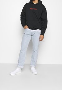 Zwarte hoodie met rode tekst, gecombineerd met lichtblauwe en witte gestreepte broek. Model draagt witte sneakers met groene accenten.