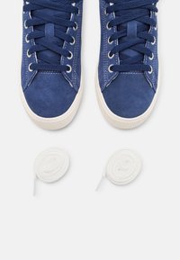 Blå mockaskor med vita gummisulor och snördesign, kompletterade med två vita runda skosnören.