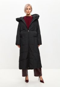 Černý prošívaný kabát s kapucí, který má plný přední zip, dvě velké kapsy a dlouhý, splývavý design. Hladká textura a podšívkový vzor.