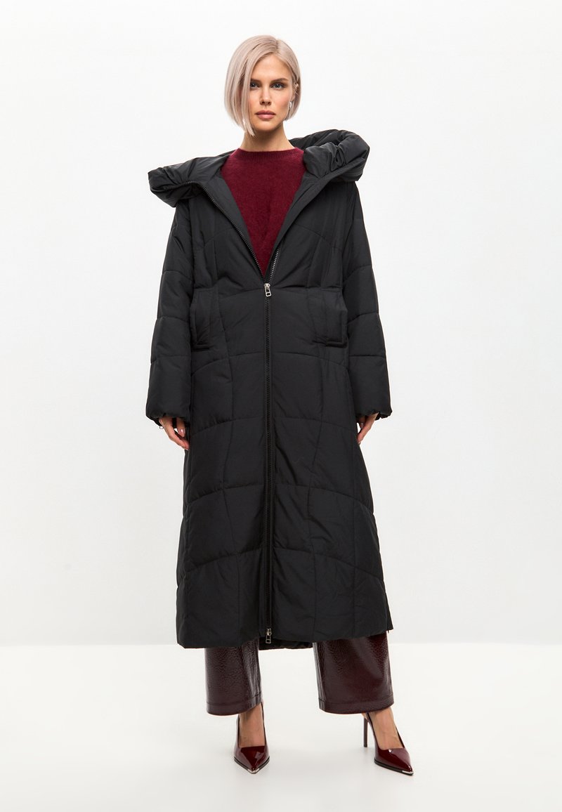 Černý prošívaný kabát s kapucí, který má plný přední zip, dvě velké kapsy a dlouhý, splývavý design. Hladká textura a podšívkový vzor.