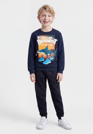 Garçon souriant portant un sweat-shirt bleu marine avec un motif requin et du texte, pantalon noir et baskets blanches sur un fond blanc uni.