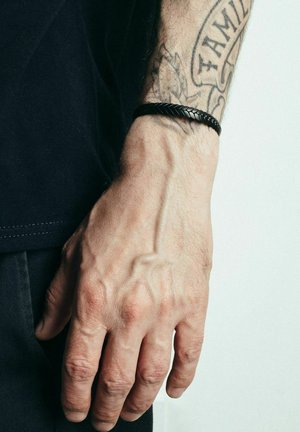 Schwarzes geflochtenes Lederarmband, das an einer Hand getragen wird und das strukturierte Design hervorhebt. Das Handgelenk zeigt sichtbare Venen und ein Tattoo.