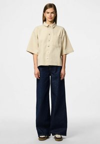 Svetlo bež srajca s kratkimi rokavi in gumbom, z ovratnikom in žepom na prsih, v kombinaciji s širokimi temno modrimi kavbojkami in športnimi copati.