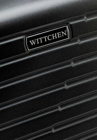 Detalle de maleta rígida negra con ranuras horizontales y una placa rectangular plateada con la marca "WITTCHEN" centrada en la superficie.