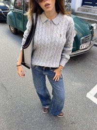 Pelēks džemperis ar adījumu un apkakli, kombinēts ar platām zilām džinsa biksēm un brūnām kurpēm. Pār vienu plecu karājas melna soma.