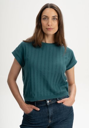 Femme aux longs cheveux bruns portant un pull à manches courtes turquoise et un jean bleu foncé, debout les mains dans les poches devant un fond uni.