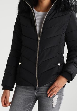 Žena v černé prošívané bundě s kožešinovou kapucí, stříbrným zipem a kapsami na druky, v kombinaci s roztrhanými šedými džíny a bílým topem.