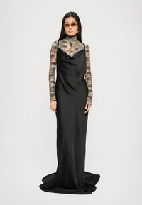 Vrouw in een lange zwarte satijnen jurk over een bloemenprint sheer lange mouwen top, met oranje-tinten zonnebril en gouden oorringen.