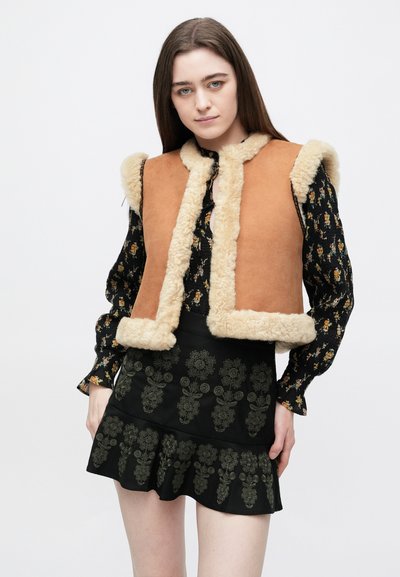 Καφέ σουέντ γιλέκο με κρέμα επένδυση από συνθετική γούνα· σχεδιασμός χωρίς γιακά. Το μοντέλο φοράει μια φλοράλ μπλούζα και μια μαύρη φούστα με περίπλοκα πράσ�ινα σχέδια.