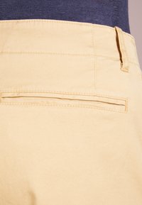 Yellow cotton pants with a smooth texture, featuring a rear pocket with a thin stitch detail and a waistband loop for securing a belt.