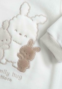 Weißer Stoff mit plüschiger Stickerei von zwei Hasen in Beigetönen. Enthält weiche Textur und Bündchendetails. Der Text lautet "Familienumarmung."