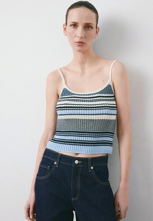 Ženska, ki nosi brezrokavnik s črtastim pletenim zgornjim delom v modri, beli in črni barvi ter temne denim hlače, stoji ob belem zidu.