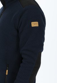 Giacca in pile navy con accenti neri sulle spalle, tessuto testurizzato e una tasca con zip che presenta un'etichetta beige con il logo "Swedemount".